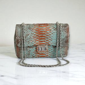 Op de afbeelding: Een rechthoekige handtas met een zilveren kettingriem. De tas heeft een slangenpatroon in tinten van teal, bruin en oranje. De sluiting is zilver. Een modieus accessoire.