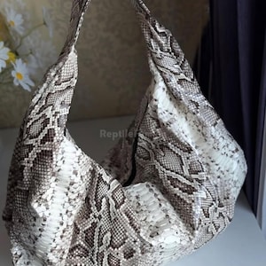 Peut inclure: Un grand sac à main à motif peau de serpent blanc cassé et marron, avec une seule bandoulière. Le sac a un design souple, de style sac hobo. Le texte "Reptile" est visible sur le sac.