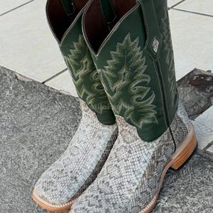 Puede incluir: Un par de botas de vaquero con un estampado de piel de serpiente en el pie y una parte superior de cuero verde. Las botas tienen una puntera cuadrada y una suela marrón claro. La parte superior presenta costuras decorativas.