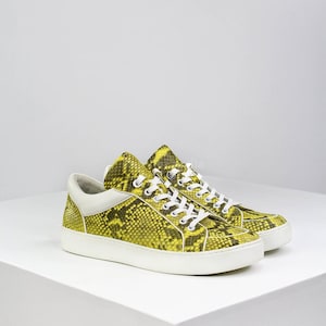 May include: A pair of yellow and black snake-print sneakers with white laces and a white sole. The sneakers have a low-top design with a white heel counter. The shoes are displayed on a white surface.