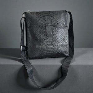 May include: Black leather crossbody bag with a textured snakeskin pattern. The bag features a flap closure, a front pocket, and an adjustable shoulder strap. The bag is displayed on a dark gray surface.