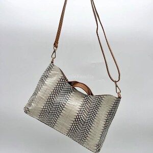 Op de afbeelding: Een handtas met wit en zwart patroon met een bruine handgreep en schouderriem. De tas heeft een rechthoekige vorm en wordt opgehangen door een bruine riem. Het patroon van de tas lijkt op slangenleer.