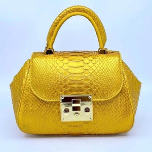 May include: A small, structured handbag in a vibrant gold colour. The bag features a textured, snakeskin-like pattern and a top handle. A gold-toned clasp secures the flap closure. The bag's design is compact and stylish, suitable for various occasions.