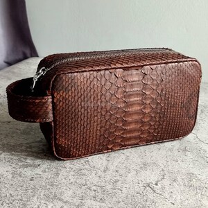 May include: A brown, rectangular travel bag with a textured snakeskin pattern. The bag has a silver zipper closure and a small handle on one side. The bag is approximately 9 inches long, 5 inches wide, and 5 inches tall.