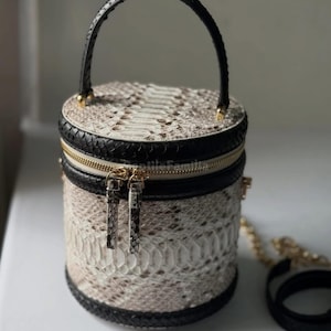 Könnte beinhalten: Zylindrische Handtasche mit Schlangenmuster in Beige und Weiß, akzentuiert mit schwarzem Lederbesatz und einem schwarzen Griff. Verfügt über einen goldenen Reißverschluss und einen abnehmbaren goldenen Kettenriemen.