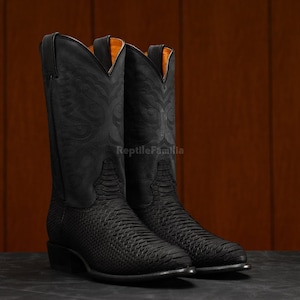 Könnte beinhalten: Ein Paar schwarze Cowboy-Stiefel mit geprägten Mustern auf dem Schaft. Die Stiefel haben einen strukturierten Vamp und eine spitze Zehenpartie. Das Innenfutter ist orange.