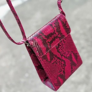 Könnte beinhalten: Eine kleine, rechteckige Umhängetasche in leuchtendem Fuchsia mit Schlangenmuster. Die Tasche hat einen Klappverschluss und einen passenden Riemen. Das Schlangenmuster weist schwarze und dunkelrosa Schuppen auf.