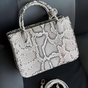 Peut inclure: Un sac à main rectangulaire avec un motif peau de serpent dans les tons gris et blanc. Le sac a une poignée supérieure et une bandoulière amovible. La quincaillerie est de couleur dorée.