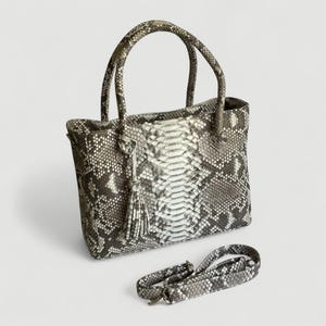 Pode incluir: Uma mala tote de pele de cobra com alças a condizer e uma alça de ombro removível. A mala apresenta uma borla e um padrão de escamas cinzentas, brancas e pretas. A mala é retangular e tem uma forma estruturada.
