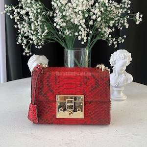 Könnte beinhalten: Rote Crossbody-Tasche aus Schlangenleder mit goldfarbenem Verschluss und Kettenriemen. Die Tasche hat ein strukturiertes Muster und einen kleinen Klappenverschluss. Im Hintergrund weiße Blumen in einer Glasvase und eine weiße Büste.