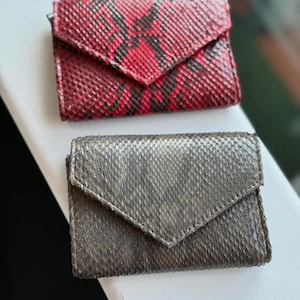 Może przedstawiać: Dwa małe, prostokątne portfele z klapkami. Górny portfel jest czerwony z czarnym wzorem skóry węża. Dolny portfel jest szary ze wzorem skóry węża. Oba portfele wykonane są z teksturowanego materiału.