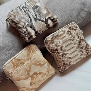 Puede incluir: Tres monederos cuadrados de piel de serpiente. Los monederos son de color marrón, beige y crema, con cierre de cremallera. El patrón de piel de serpiente es visible en cada monedero, con variaciones de tamaño y color.