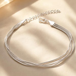 Könnte beinhalten: Ein silberfarbenes mehrsträngiges Armband, bestehend aus drei feinen Schlangenkettchen, die miteinander verflochten sind. Das Armband verfügt über einen polierten Metallverschluss und eine verstellbare Verlängerungskette mit runden Gliedern.