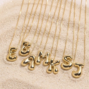 Puede incluir: Collares con iniciales doradas sobre una superficie arenosa. Cada collar presenta una letra diferente del alfabeto, incluyendo E, B, A, M, K, S y J. Las letras tienen un diseño redondeado en forma de burbuja.