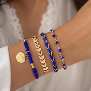 Pode incluir: Quatro pulseiras em ouro e azul são exibidas em um pulso. As pulseiras incluem uma pulseira de contas azuis com um pingente de concha dourada, uma pulseira dourada com design de chevron, uma pulseira trançada azul e uma pulseira de corrente dourada com pedras azuis.