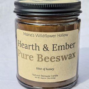 May include: A glass jar candle with a black lid and a label that reads "Hearth & Ember Pure Beeswax." The label also states "Hint of honey" and "Natural Beeswax Candle." The net weight is approximately 7.3oz (208g).