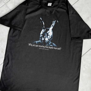 May include: Black t-shirt featuring a graphic of a figure in a rabbit mask with the text "Why are you wearing that stupid man suit?" and "Donnie Darko" in white font. The shirt is made of a soft material.