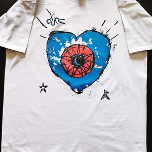 May include: White t-shirt featuring a blue heart design with a red eye in the center. The shirt has black accents and the word "cure" in a stylized font. Additional small black graphic elements are scattered around the design.