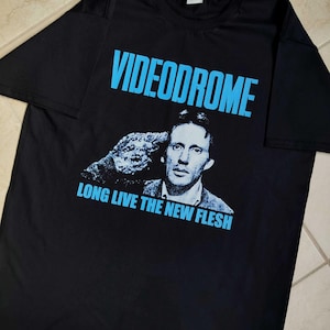 Peut inclure: T-shirt noir avec le mot "VIDEODROME" en bleu en haut d'une image en noir et blanc d'un homme. En dessous, le texte "LONG LIVE THE NEW FLESH" est imprimé en bleu.