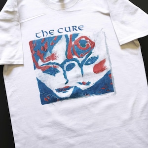 Könnte beinhalten: Weißes T-Shirt mit dem Bandnamen "The Cure" in blauer Schrift über einer quadratischen Grafik. Die Grafik zeigt zwei Gesichter und eine Rose in roten und blauen Pinselstrichen.