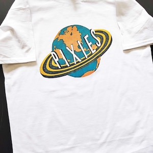 Puede incluir: Una camiseta blanca de manga corta con un estampado gráfico de un planeta con anillos amarillos. La palabra PIXIES está impresa en letras mayúsculas blancas en el centro del planeta, coloreado en tonos azules y naranjas.