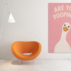 Puede incluir: Un interior moderno con una silla naranja, una l&aacute;mpara de pie blanca y una obra de arte rosa. La obra muestra un ganso de dibujos animados con el texto "ARE YOU POOPING?" en blanco. La habitaci&oacute;n tiene un dise&ntilde;o minimalista.