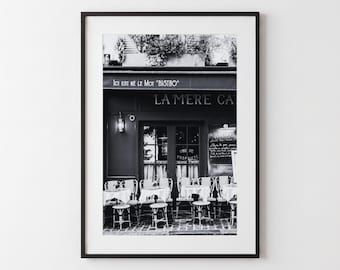 May include: A black and white framed wall art print depicting a Parisian bistro exterior. The storefront features the text 'ICI EST NE LE MOT BISTRO' and 'LA MERE CATHERINE' above a window, with outdoor tables and chairs arranged on the sidewalk.