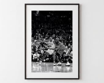 Op de afbeelding: Een zwart-wit ingelijste foto van twee basketballers in actie op een basketbalveld. Eén speler draagt een shirt met het woord Lakers zichtbaar, terwijl de andere speler verdedigt. De scène is vastgelegd tijdens een wedstrijd met publiek op de achtergrond.