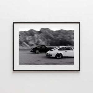 May include: A black-and-white framed wall art print featuring a motion-blurred action shot of a black sports car and a white sports car driving side-by-side on a road with a rocky landscape in the background, presented in a simple black frame.