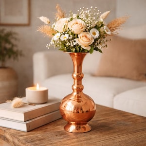 May include: A copper-colored vase with a bulbous base and a flared top holds a bouquet of cream-colored roses, daisies, and dried grasses. A lit candle and stacked books sit on a wooden table, creating a warm, inviting scene.