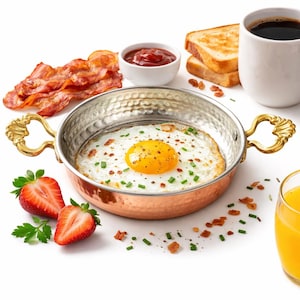 May include: A breakfast spread featuring a fried egg in a copper pan with gold handles, crispy bacon, toast, coffee, ketchup, strawberries, and orange juice. The egg has a bright yellow yolk and is sprinkled with chives.
