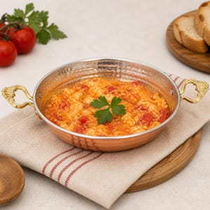 May include: A copper-colored pan filled with scrambled eggs and diced tomatoes, garnished with a sprig of parsley. The pan has decorative gold-colored handles and sits on a beige linen napkin. Sliced bread and fresh tomatoes are in the background.