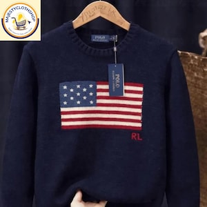 Può includere: Maglione blu navy con un disegno della bandiera americana sul davanti. La bandiera presenta stelle bianche su sfondo blu e strisce rosse e bianche. Il maglione ha un girocollo e maniche lunghe. L'etichetta del marchio dice "POLO".