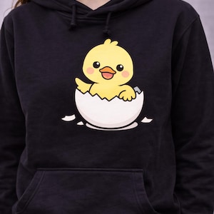May include: Black hoodie with a cartoon yellow chick emerging from a white egg. The chick has rosy cheeks and is waving. The text "5 x 7 Hoop" is at the top. The bottom of the hoodie has the text "DST, PES, JEF, EXP, HUS, VP3, XXX".