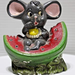 May include: A vintage ceramic figurine of a gray mouse seated in a watermelon slice. The mouse has large ears with pink interiors, holding a yellow fruit. The watermelon is red with black seeds and a green rind.