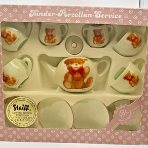 May include: A boxed porcelain tea set featuring a teddy bear design. The set includes a teapot, cups, saucers, and a sugar bowl. The box is pink with white floral patterns and the text "Kinder-Porzellan-Service". The set is a limited edition collector's item.
