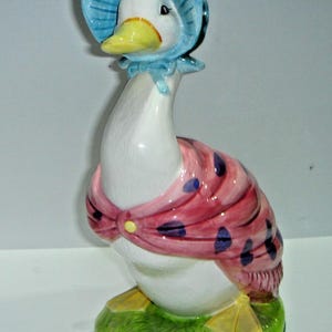 May include: A ceramic goose figurine, painted white with a yellow beak and feet. It wears a blue bonnet and a pink scarf with blue polka dots. The goose stands on a green base, a charming decorative item.