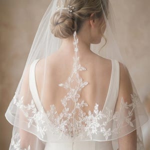 May include: A bridal veil with intricate floral lace detailing. The sheer, white veil cascades over the shoulders, showcasing a delicate pattern of leaves and flowers. A hair comb with pearls secures the veil to the updo.
