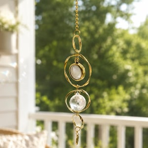 May include: A hanging suncatcher featuring a gold-toned metal chain with interconnected twisted circular rings. The design includes a smooth, opaque white stone set in a gold bezel and a clear, multifaceted crystal sphere suspended within the rings.