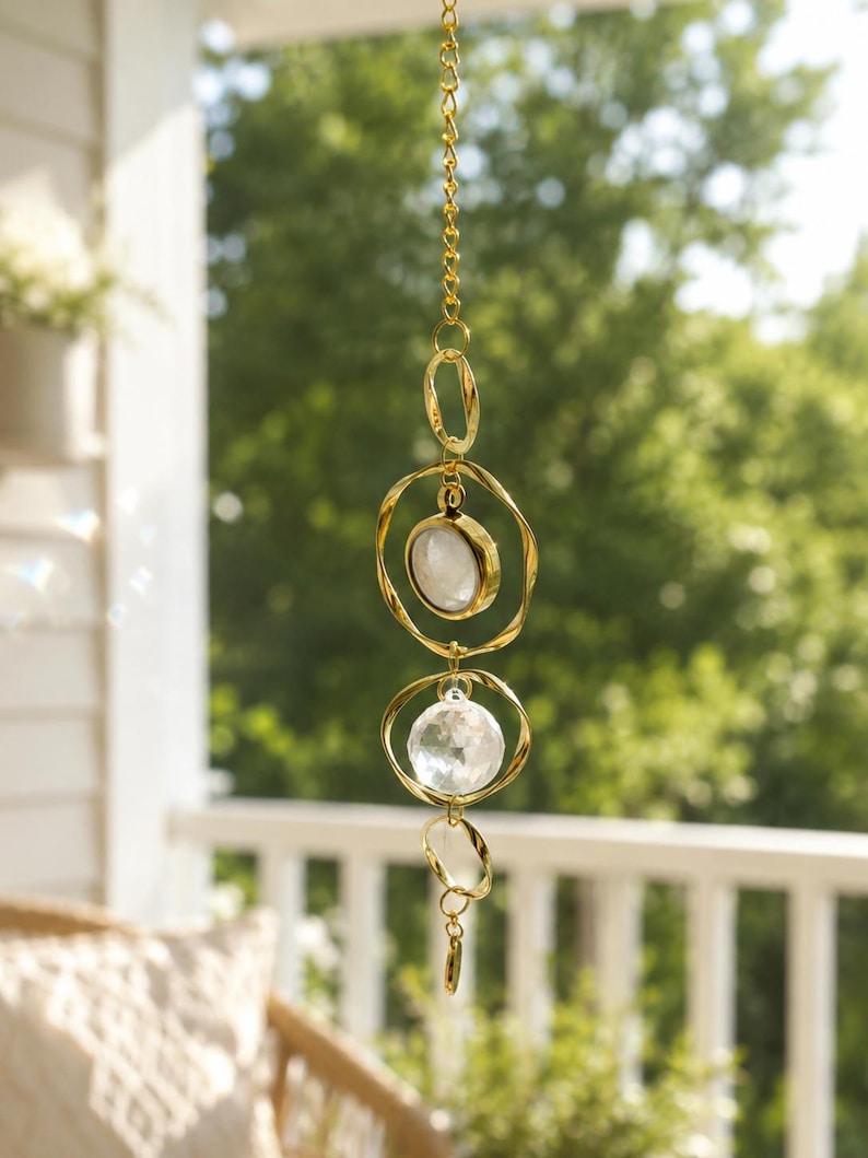 May include: A hanging suncatcher featuring a gold-toned metal chain with interconnected twisted circular rings. The design includes a smooth, opaque white stone set in a gold bezel and a clear, multifaceted crystal sphere suspended within the rings.