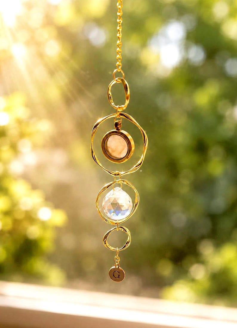 May include: A gold-toned hanging ornament featuring a vertical chain with three irregular circular rings. The top ring holds a smooth, pale pink circular stone, the middle ring contains a faceted clear crystal, and a small round charm with the letter G hangs below.