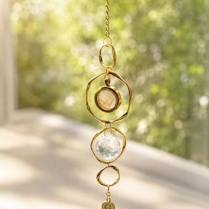 May include: A gold-toned hanging ornament featuring a vertical chain with three irregular circular metal rings. The top ring holds a circular stone, the middle ring contains a faceted clear crystal prism, and a small round charm with the letter G hangs below.