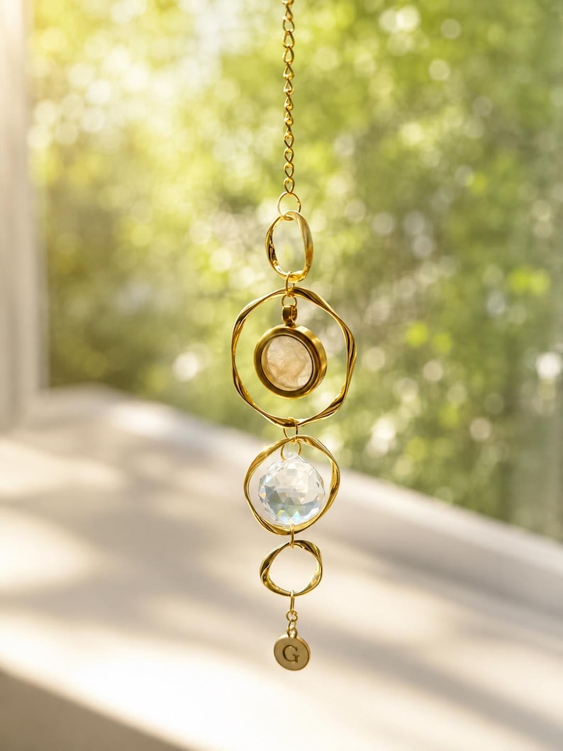May include: A gold-toned hanging ornament featuring a vertical chain with three irregular circular metal rings. The top ring holds a circular stone, the middle ring contains a faceted clear crystal prism, and a small round charm with the letter G hangs below.