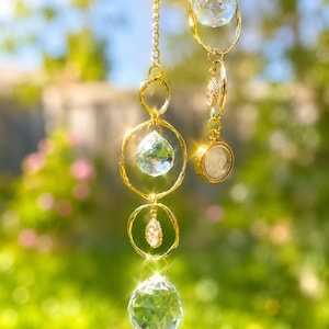May include: A gold-coloured hanging crystal suncatcher with multiple clear, faceted crystal orbs and decorative accents. The suncatcher is suspended from a delicate chain and features a variety of shapes and sizes, reflecting light. The background is a soft, blurred green.