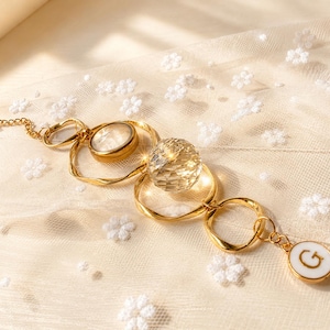 May include: A gold-toned chain necklace featuring a series of irregular circular links, a smooth round stone, a faceted clear crystal sphere, and a small white circular pendant with the letter G printed in black, all connected in a linear arrangement.