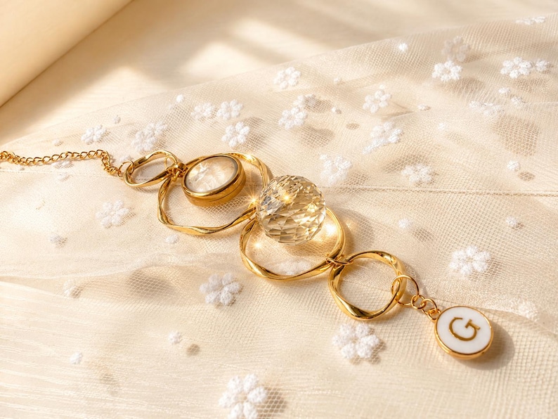 May include: A gold-toned chain necklace featuring a series of irregular circular links, a smooth round stone, a faceted clear crystal sphere, and a small white circular pendant with the letter G printed in black, all connected in a linear arrangement.