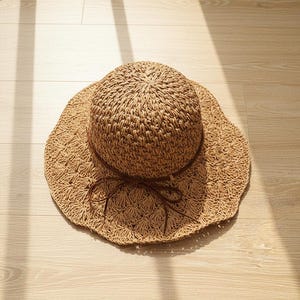 May include: A woven straw sun hat with a wide, scalloped brim and a rounded crown. The hat is tan with a brown band tied in a bow. The hat is designed for sun protection and casual wear.