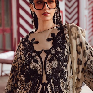 Puede incluir: Una mujer con gafas de sol cuadradas grandes y pendientes largos. Lleva un vestido beige y negro estampado con bordados florales negros. Una bufanda transparente con estampado de leopardo está drapeada sobre su hombro.