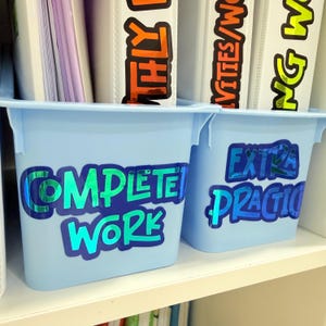 May include: Two light blue plastic bins with labels that read "COMPLETE WORK" and "EXTRA PRACTICE" in a shiny, iridescent font. The bins are on a white shelf, with binders visible in the background.