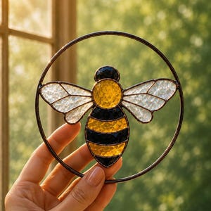 May include: A circular stained glass suncatcher featuring a central bee design. The bee has a yellow textured glass thorax and abdomen with black glass stripes. The wings are made of clear, iridescent textured glass, all encased in a dark metal frame.